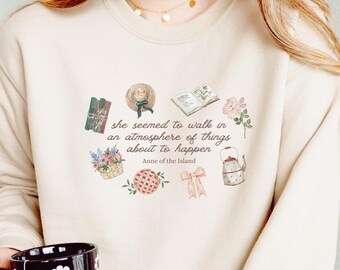 May include: Beige sweatshirt with a floral and text design. The design includes a quote from "Anne of the Island" and illustrations of a hat, flowers, and a pie. A black mug with white daisy patterns is held in the hand.