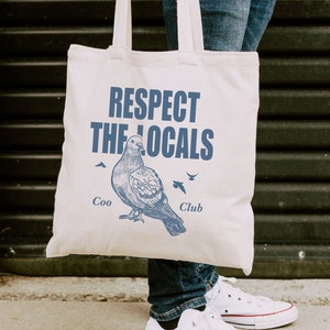 May include: A white canvas tote bag with the text "Respect the Locals" in blue lettering. A blue illustration of a pigeon with the text "Coo Club" is below the main text.
