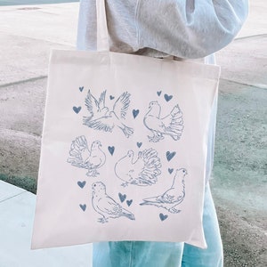 May include: A white canvas tote bag with a blue outline of doves and hearts. The doves are in various poses, some flying, some perched, and some with their wings spread. The bag is being held by a person wearing a grey sweatshirt.