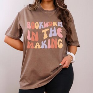 Puede incluir: Una camiseta marrón con el texto "BOOKWORM IN THE MAKING" en letras de colores de estilo retro.