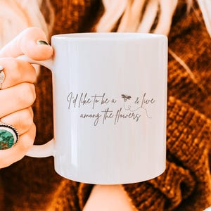 Peut inclure: Mug en céramique blanche avec le texte "I'd like to be a bee & live among the flowers" dans un style manuscrit. La tasse a une anse et est tenue par une personne. L'arrière-plan est flou.