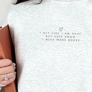 Puede incluir: Una sudadera gris con el texto "I act like I am okay but deep down I need more books" y un pequeño símbolo de corazón.