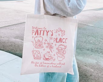 Puede incluir: Bolso tote de color crema con una ilustración y texto en rosa. El texto dice "Welcome to PATTY'S PLACE" con dibujos de una tetera, flores, libros y tazas de té. El texto inferior dice "the fun of homemaking without the bother of a husband."