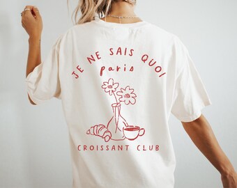 Puede incluir: Camiseta color crema con un gráfico rojo en la espalda. El gráfico incluye el texto "JE NE SAIS QUOI paris" encima de un jarrón de flores, un cruasán y una taza de café. El texto "CROISSANT CLUB" está debajo.