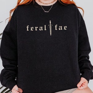 Puede incluir: Sudadera negra con el texto "feral fae" en fuente gótica, con un gráfico de espada. La parte superior de manga larga es de color oscuro y parece estar hecha de un material suave. El diseño es de color marrón claro.
