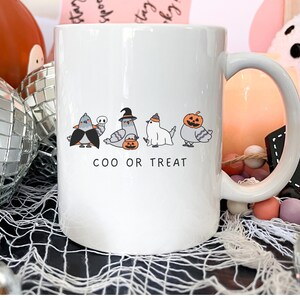 May include: White ceramic mug with a Halloween-themed design. The mug features cartoon pigeons dressed in costumes, with the text "COO OR TREAT". The design includes a bat, a skull, a witch, and a pumpkin. The mug has a large handle.