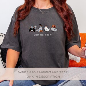 K&ouml;nnte beinhalten: Dunkelgraues Comfort Colors T-Shirt mit einem Halloween-Motiv. Das Shirt zeigt eine Reihe von Cartoon-Tauben in Kost&uuml;men mit dem Text "COO OR TREAT" darunter. Eine schwarze Tasse mit Geisterdesign wird in der Hand gehalten.