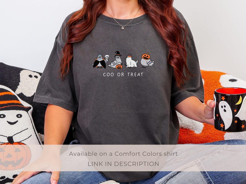 K&ouml;nnte beinhalten: Dunkelgraues Comfort Colors T-Shirt mit einem Halloween-Motiv. Das Shirt zeigt eine Reihe von Cartoon-Tauben in Kost&uuml;men mit dem Text "COO OR TREAT" darunter. Eine schwarze Tasse mit Geisterdesign wird in der Hand gehalten.