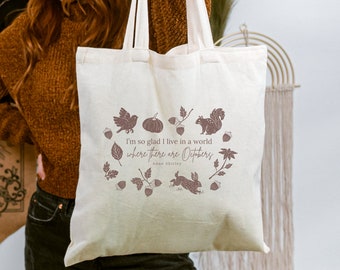 Puede incluir: Una bolsa de lona blanca con una impresión marrón de una cita de Anne Shirley: "Estoy tan contenta de vivir en un mundo donde hay Octobres." La impresión también incluye ilustraciones de hojas, bellotas, una ardilla, un pájaro y una calabaza.