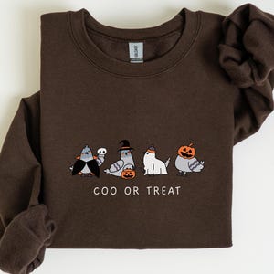 K&ouml;nnte beinhalten: Ein dunkelbraunes Sweatshirt mit einem Halloween-Motiv. Die Grafik zeigt vier Cartoon-Tauben in Kost&uuml;men, darunter der Text "COO OR TREAT". Die Tauben sind als Vampir, Hexe, Geist und K&uuml;rbis verkleidet.