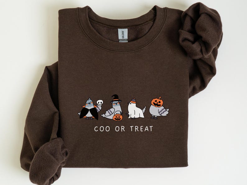 K&ouml;nnte beinhalten: Ein dunkelbraunes Sweatshirt mit einem Halloween-Motiv. Die Grafik zeigt vier Cartoon-Tauben in Kost&uuml;men, darunter der Text "COO OR TREAT". Die Tauben sind als Vampir, Hexe, Geist und K&uuml;rbis verkleidet.