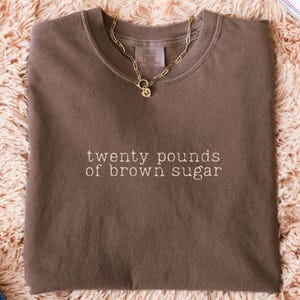 Pode incluir: Uma camiseta marrom com o texto "twenty pounds of brown sugar" em fonte marrom claro. Um colar de corrente dourada com um pequeno pingente está sobre a camiseta. A camiseta está dobrada sobre uma superfície fofa marrom claro.