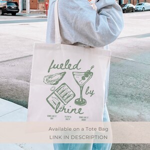 Puede incluir: Una bolsa de tela blanca con una ilustraci&oacute;n verde de una copa de martini, ostras y una lata de sardinas. El texto "fueled by brine" est&aacute; escrito en cursiva verde. El texto "SOME LIKE IT DRY" est&aacute; escrito en verde m&aacute;s peque&ntilde;o debajo de la ilustraci&oacute;n. El texto "SOME LIKE IT BRINY" est&aacute; escrito en verde m&aacute;s peque&ntilde;o debajo de la ilustraci&oacute;n. El texto "FILTHY & SALTY" est&aacute; escrito en verde m&aacute;s peque&ntilde;o debajo de la ilustraci&oacute;n. El texto "Available on a Tote Bag" est&aacute; escrito en negro debajo de la ilustraci&oacute;n. El texto "LINK IN DESCRIPTION" est&aacute; escrito en negro debajo de la ilustraci&oacute;n.