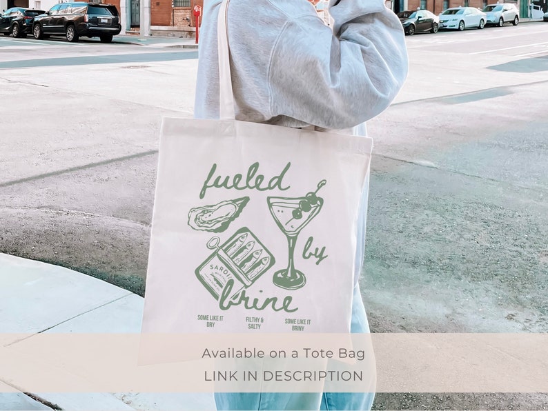 Puede incluir: Una bolsa de tela blanca con una ilustraci&oacute;n verde de una copa de martini, ostras y una lata de sardinas. El texto "fueled by brine" est&aacute; escrito en cursiva verde. El texto "SOME LIKE IT DRY" est&aacute; escrito en verde m&aacute;s peque&ntilde;o debajo de la ilustraci&oacute;n. El texto "SOME LIKE IT BRINY" est&aacute; escrito en verde m&aacute;s peque&ntilde;o debajo de la ilustraci&oacute;n. El texto "FILTHY & SALTY" est&aacute; escrito en verde m&aacute;s peque&ntilde;o debajo de la ilustraci&oacute;n. El texto "Available on a Tote Bag" est&aacute; escrito en negro debajo de la ilustraci&oacute;n. El texto "LINK IN DESCRIPTION" est&aacute; escrito en negro debajo de la ilustraci&oacute;n.