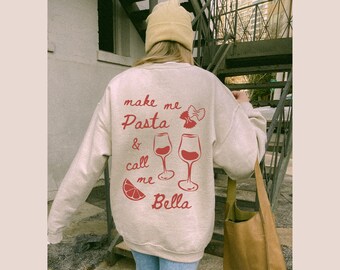 Peut inclure: Un sweat-shirt beige avec un motif rouge qui dit "make me pasta & call me bella" avec un verre de vin rouge, des pâtes rouges et une tranche d'orange rouge.