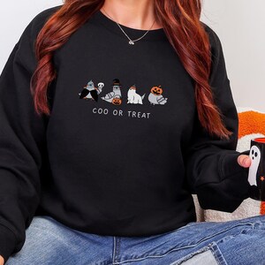 K&ouml;nnte beinhalten: Schwarzes Sweatshirt mit einem Halloween-Motiv. Das Design zeigt f&uuml;nf Cartoon-Tauben in Kost&uuml;men, darunter der Text "COO OR TREAT". Eine schwarze Tasse mit Geister- und Sterndesigns wird gehalten.