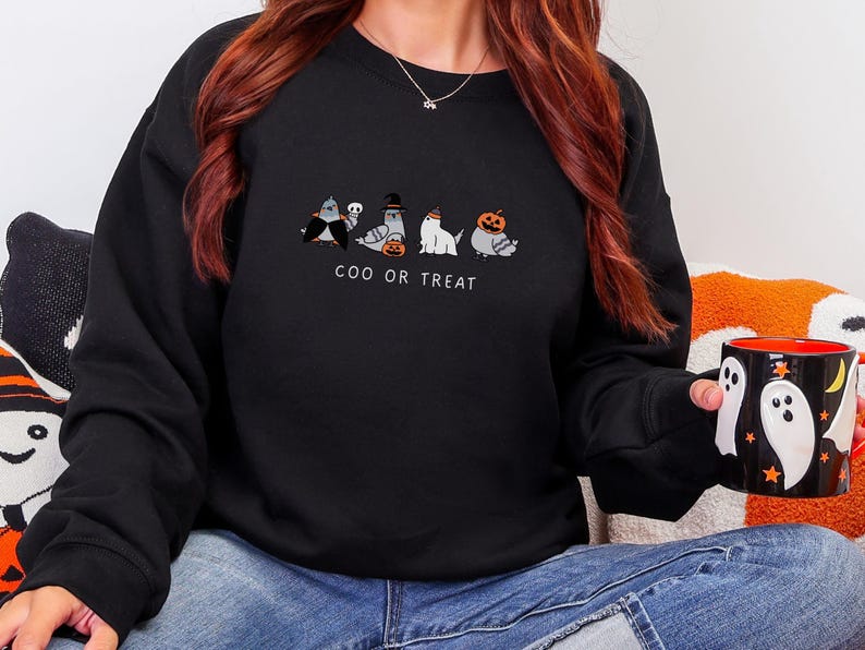 K&ouml;nnte beinhalten: Schwarzes Sweatshirt mit einem Halloween-Motiv. Das Design zeigt f&uuml;nf Cartoon-Tauben in Kost&uuml;men, darunter der Text "COO OR TREAT". Eine schwarze Tasse mit Geister- und Sterndesigns wird gehalten.
