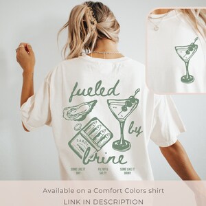 Puede incluir: Una camiseta blanca con un dise&ntilde;o gr&aacute;fico verde que dice "fueled by brine" e incluye una ostra, una lata de sardinas y una copa de martini con aceitunas.