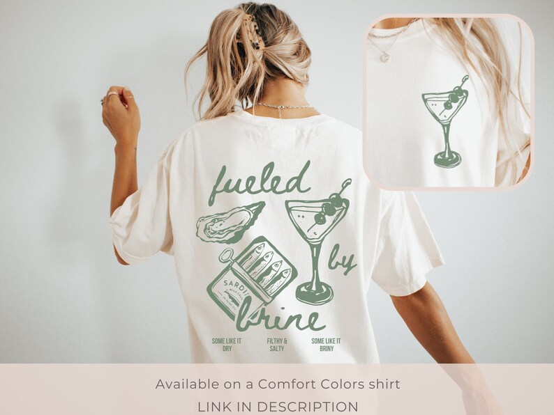 Puede incluir: Una camiseta blanca con un dise&ntilde;o gr&aacute;fico verde que dice "fueled by brine" e incluye una ostra, una lata de sardinas y una copa de martini con aceitunas.