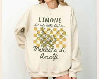Peut inclure: Sweat-shirt crème avec un motif graphique. Le motif comprend le texte "LIMONE dal sole della Costiera" et "Mercato di Amalfi". Le motif comprend un citron, un sac de courses et un motif à carreaux verts et jaunes.