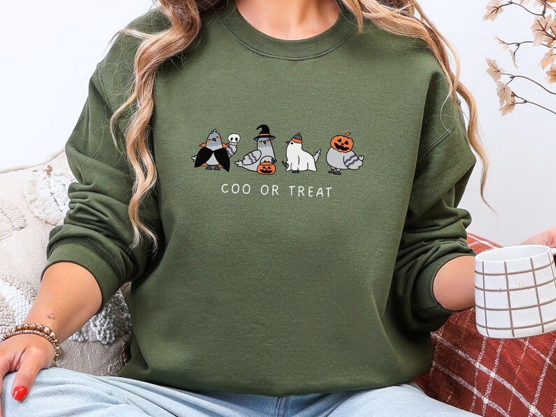 K&ouml;nnte beinhalten: Olivgr&uuml;nes Sweatshirt mit einem Halloween-Motiv. Das Design zeigt Cartoon-Tauben in Kost&uuml;men, darunter ein Vampir und eine Hexe, mit dem Text "COO OR TREAT" darunter. Das Sweatshirt ist ein l&auml;ssiger Stil.