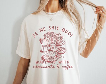 Puede incluir: Una camiseta blanca con un estampado gráfico rojo de una cafetera, croissants y el texto "Je ne sais quoi Wake me with croissants & coffee".