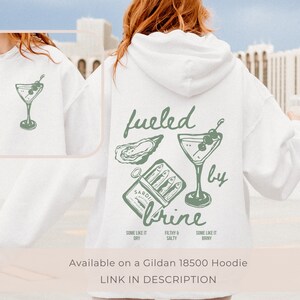 Puede incluir: Sudadera blanca con un dise&ntilde;o gr&aacute;fico verde que presenta una copa de martini, una ostra y una lata de sardinas con el texto "fueled by brine". El texto debajo del gr&aacute;fico dice "Some like it dry, Filthy & Salty, Some like it briny".