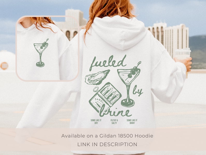 Puede incluir: Sudadera blanca con un dise&ntilde;o gr&aacute;fico verde que presenta una copa de martini, una ostra y una lata de sardinas con el texto "fueled by brine". El texto debajo del gr&aacute;fico dice "Some like it dry, Filthy & Salty, Some like it briny".