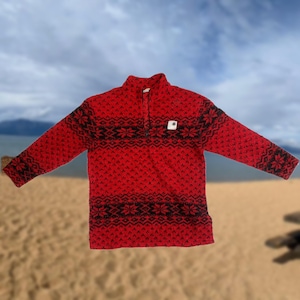 May include: Red fleece pullover with a black and white geometric pattern. The pullover has a zipper at the neck and a small logo patch on the left chest.
