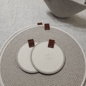 May include: A woven grey and white rope basket with matching placemats and coasters. The basket has a brown leather handle. The coasters and placemats are white with grey rope trim. The coasters have brown leather tags.