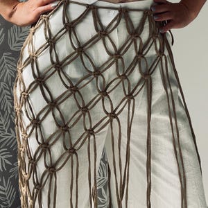 Puede incluir: Un pantalón blanco con una superposición de macramé marrón. El macramé está anudado en un patrón geométrico, creando una franja decorativa a lo largo de las piernas. El pantalón está hecho de una tela de color claro.