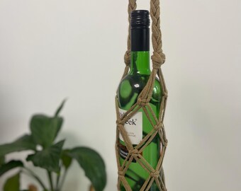 Puede incluir: Una botella de vino verde en un colgador de plantas de macramé. La botella tiene una tapa negra y una etiqueta. El macramé está hecho de cuerda marrón y tiene una borla. Una planta verde en una maceta está en el fondo.