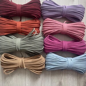 May include: A selection of macrame cords in a variety of colours, such as burgundy, orange, sage green, tan, lavender, pink, fuchsia, and light blue. Each cord is neatly coiled and tied, demonstrating the range of colours available for crafting projects.
