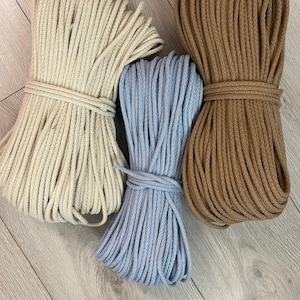 Könnte beinhalten: Drei Bündel Baumwollseil in Creme, Hellblau und Braun. Jedes Bündel ist fest gewickelt und mit passendem Seil gesichert. Die Seile sind auf einer hellen Holzmaserung angeordnet.