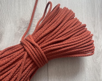 May include: Coiled orange-red macrame rope, tightly wound and secured with a wrapped section. The rope is thick and appears to be made of a soft, textured material. The bundle is shown against a light gray wood grain background.