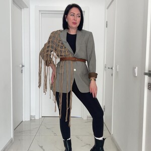Puede incluir: Una blazer a rayas grises, cinturón marrón y chal de macramé beige. El atuendo incluye leggings negros y botas negras. La blazer tiene un solo botón y puños marrones. El chal de macramé tiene flecos largos.
