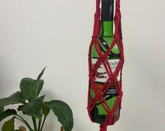 Puede incluir: Un portabotellas de vino de macramé rojo con borla, que sostiene una botella de vino verde. La botella tiene una tapa negra y una etiqueta con texto. Una planta en maceta está en el fondo.