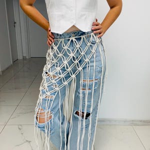 May include: A white macrame skirt with a geometric pattern is worn over light blue distressed jeans. The skirt has long fringe and is paired with a white vest.