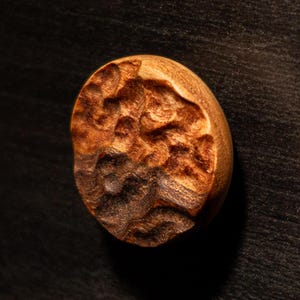 Op de afbeelding: Een close-up van een houten kastknop met een textuur, organisch ontwerp. De knop heeft een warme bruine kleur en lijkt met de hand gesneden te zijn. De achtergrond is een donker oppervlak met houtnerf.