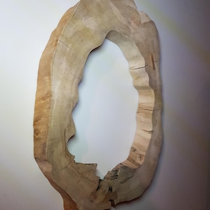 Può includere: Un elemento decorativo da parete di forma ovale in legno naturale. Il legno è di colore marrone chiaro con venature e crepe visibili. Lo spazio interno è aperto, creando un aspetto simile a una cornice. Il pezzo è montato su una parete bianca.