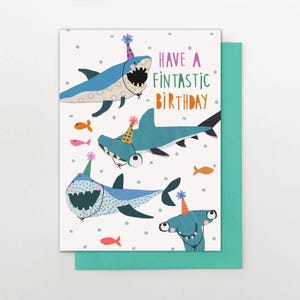 May include: A birthday card with a teal envelope features cartoon sharks wearing party hats. The card reads "Have a Fintastic Birthday" with colourful text and includes illustrations of fish and polka dots.