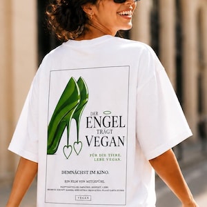Op de afbeelding: Wit T-shirt met een afbeelding van groene hakken en de tekst "DER ENGEL TRÄGT VEGAN" in het zwart. Het shirt bevat ook de tekst "FÜR DIE TIERE. LEBE VEGAN." en "DEMNÄCHST IM KINO."