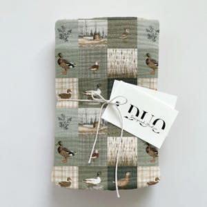 Peut inclure: Une couverture pli&eacute;e avec un motif patchwork repr&eacute;sentant des canards, des quenouilles et des paysages dans des tons de vert, de marron et de beige. Une &eacute;tiquette blanche avec le texte "DUO Designed Studio" est attach&eacute;e &agrave; la couverture avec de la ficelle.