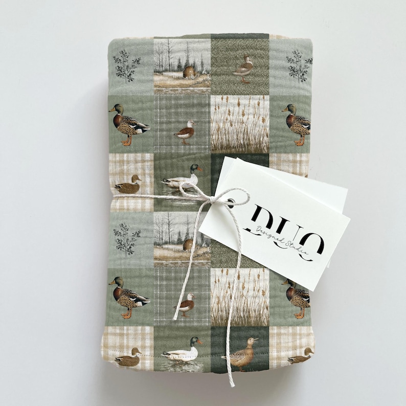 Peut inclure: Une couverture pli&eacute;e avec un motif patchwork repr&eacute;sentant des canards, des quenouilles et des paysages dans des tons de vert, de marron et de beige. Une &eacute;tiquette blanche avec le texte "DUO Designed Studio" est attach&eacute;e &agrave; la couverture avec de la ficelle.