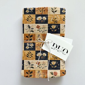 Peut inclure: Un carr&eacute; de tissu pli&eacute; avec un motif &agrave; carreaux noirs, beiges et orange. Chaque carr&eacute; pr&eacute;sente un motif floral diff&eacute;rent dans des tons de brun, blanc et rouge. Le tissu est nou&eacute; avec une ficelle blanche et une &eacute;tiquette blanche avec le texte "DUO Design Studio".