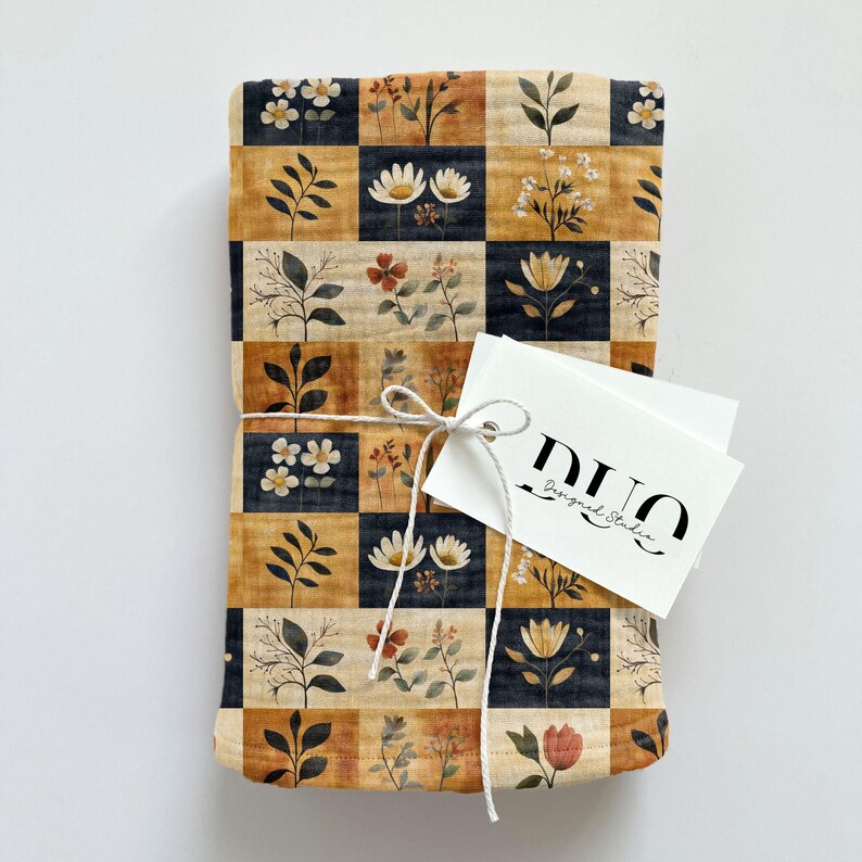 Peut inclure: Un carr&eacute; de tissu pli&eacute; avec un motif &agrave; carreaux noirs, beiges et orange. Chaque carr&eacute; pr&eacute;sente un motif floral diff&eacute;rent dans des tons de brun, blanc et rouge. Le tissu est nou&eacute; avec une ficelle blanche et une &eacute;tiquette blanche avec le texte "DUO Design Studio".