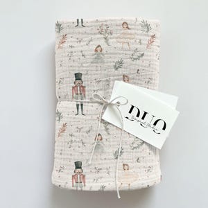Puede incluir: Una manta de muselina rosa para beb&eacute; con un estampado blanco y verde de bailarinas y cascanueces. La manta est&aacute; atada con una cuerda blanca y tiene una etiqueta blanca con el texto "DUO Designed Studio".