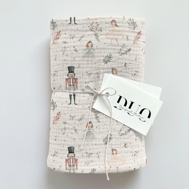 Puede incluir: Una manta de muselina rosa para beb&eacute; con un estampado blanco y verde de bailarinas y cascanueces. La manta est&aacute; atada con una cuerda blanca y tiene una etiqueta blanca con el texto "DUO Designed Studio".