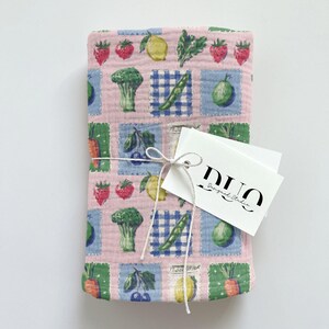 May include: A pink fabric item featuring a repeating pattern of fruits and vegetables in square blocks. The design includes strawberries, lemons, broccoli, and carrots. A white tag with the text "DUO Designed Studio" is tied to the fabric with string.