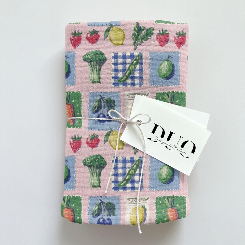 May include: A pink fabric item featuring a repeating pattern of fruits and vegetables in square blocks. The design includes strawberries, lemons, broccoli, and carrots. A white tag with the text "DUO Designed Studio" is tied to the fabric with string.